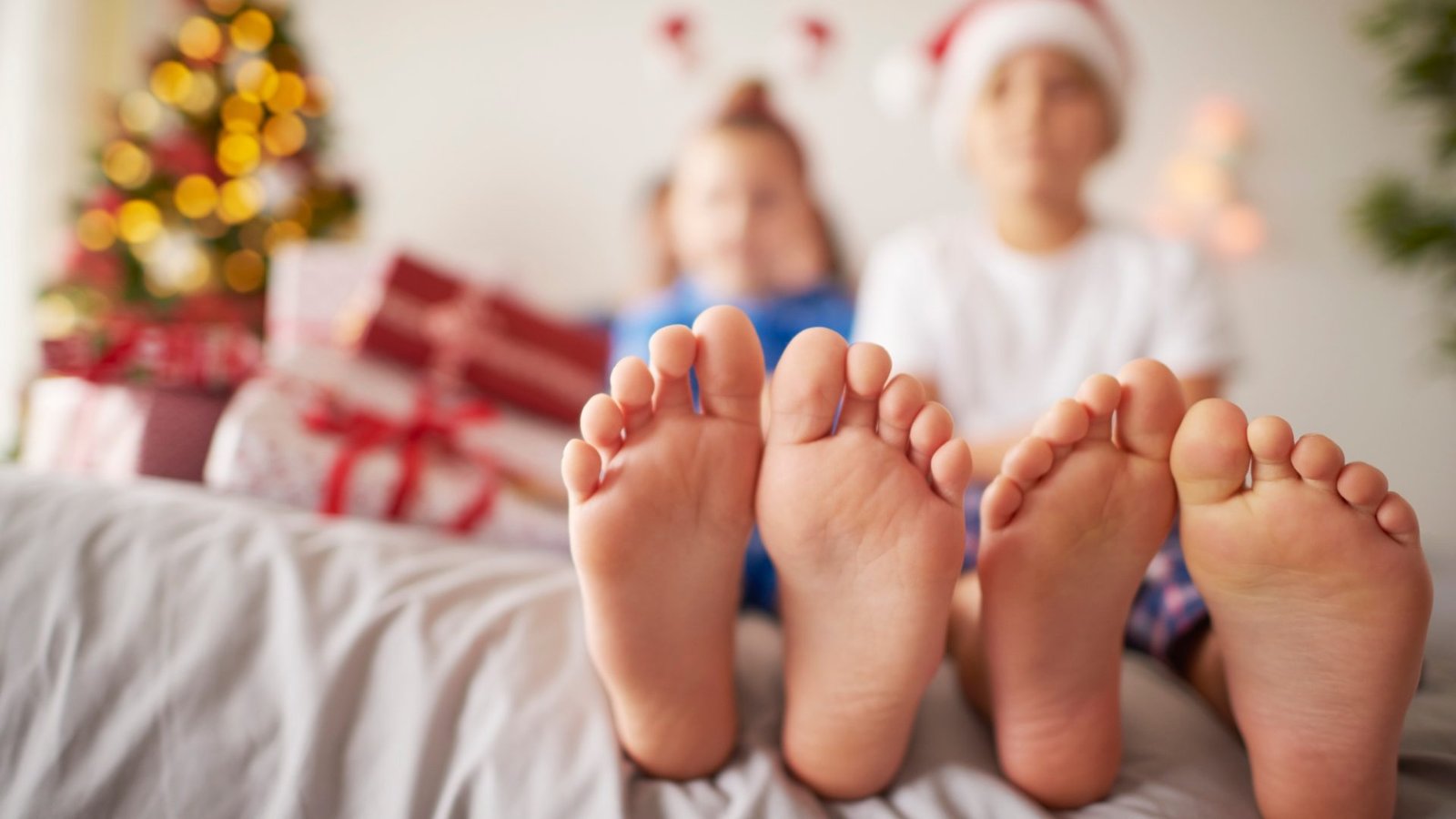 Dos niños con gorros navideños sentados en una cama, mostrando las plantas de sus pies en primer plano, con un árbol de Navidad y regalos desenfocados al fondo.