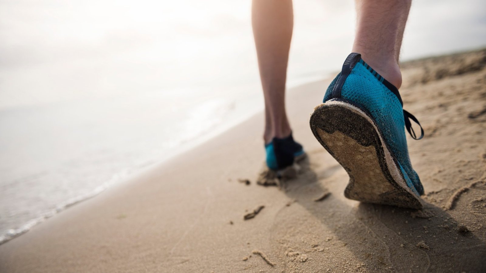 Persona caminando por la orilla de la playa con zapatillas deportivas azules, dejando huellas en la arena.